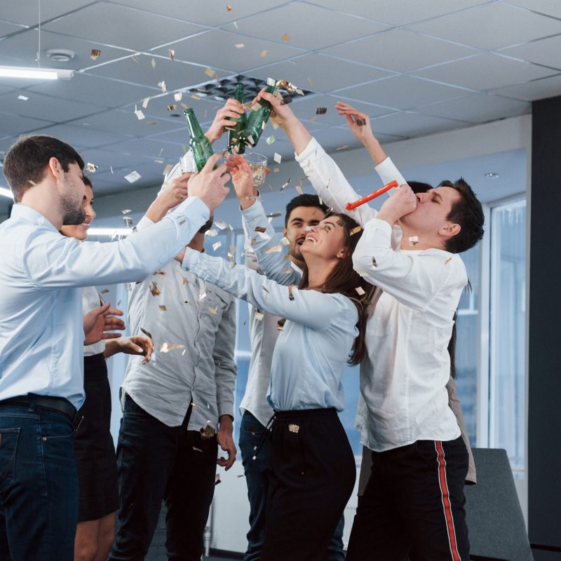 Hands up. Photo of young team in classical clothes celebrating success while holding drinks in the modern good lighted office.